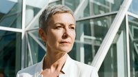 Senior woman with short gray hair and fair complexion wearing white business shirt, standing outdoors against modern glass building architecture. Corporate professional portrait.