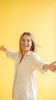 Young woman with shoulder-length blonde hair, Caucasian ethnicity, wearing white linen shirt, smiling broadly with arms outstretched against bright yellow background, expressing joy and freedom.