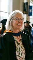 Happy senior Caucasian woman professor in graduation gown and glasses smiling at university ceremony with students in academic regalia background.
