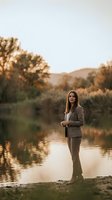 Young professional woman in beige suit standing by lake during golden hour sunset with autumn trees and mountains in background for business lifestyle.