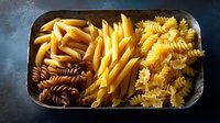 Various uncooked pasta types including penne, fusilli and farfalle arranged in rustic metal tray on dark background, raw traditional Italian macaroni assortment.