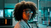 Young African American male with curly afro hairstyle wearing reflective sunglasses and light-colored shirt in modern tech office environment with neon lighting and glass partitions.