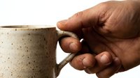 Male hand holding a rustic speckled ceramic mug, close-up shot on white background, showcasing handmade pottery craft and morning coffee culture.