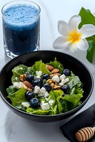 Fresh green salad with blueberries, feta cheese, mixed nuts in black bowl, blue smoothie and tropical flower on marble background. Food styling.