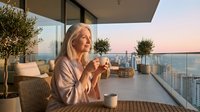 Senior Caucasian woman enjoying morning coffee on luxury apartment balcony with city skyline view during golden hour sunrise for lifestyle content.