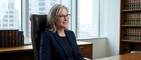 Female professional with gray hair and glasses wearing dark blazer seated at desk in modern law office with city skyline view, bookshelf background, suitable for corporate, legal, and business publications.