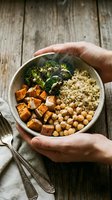 Hands holding bowl with roasted sweet potato, chickpeas, broccoli and quinoa on wooden surface. Healthy vegan meal prep and nutritious lunch bowl.