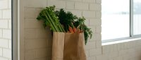 Fresh organic vegetables including carrots and leafy greens in brown paper grocery bag on kitchen counter for healthy cooking and meal preparation.
