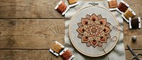 Mandala embroidery hoop with intricate floral pattern on linen fabric surrounded by colorful thread spools on rustic wooden background for crafting projects.