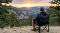 Man in camping chair watching sunrise over mountain valley with pine trees and golden sky creating peaceful outdoor adventure moment.