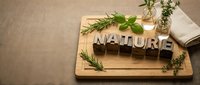 Wooden letterpress blocks spelling NATURE on a bamboo cutting board with fresh herbs, glass vials, and linen napkin on burlap background.