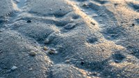 Sandy beach surface with rippled texture and golden sunlight casting shadows across fine grains, suitable for backgrounds and nature compositions.