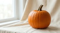 Fresh orange pumpkin on white fabric near window with soft natural light creating cozy autumn atmosphere for seasonal home decor and harvest celebrations.