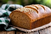 Fresh homemade whole grain bread loaf with sesame seeds on top, served on rustic wooden table with green checkered napkin, warm natural lighting.