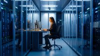 Female IT specialist working on laptop in modern server room with blue lighting and network equipment racks for technology and data center operations.