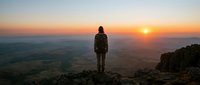 Young adult male hiker standing on mountain peak at sunrise, overlooking vast valley landscape. Outdoor adventure and exploration concept for travel and lifestyle use.