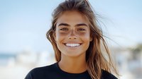 Young woman with radiant smile and windswept brown hair wearing black top against blurred beach background, natural beauty and joy captured in outdoor portrait.