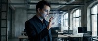 Young Caucasian businessman in dark shirt interacting with futuristic holographic interface technology in modern office workspace with industrial windows.