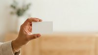 Cropped hand holding blank white card against neutral beige background with plant, suitable for business branding, contact information, and corporate identity mockups.