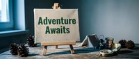 Adventure Awaits text on canvas easel with pine cones, compass, and lantern on rustic wooden table, travel and outdoor exploration concept.