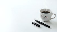 Cute coffee mug with kawaii face next to black pens on clean white desk surface for morning productivity and workspace organization concepts.