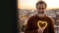 Middle-aged Caucasian man with beard holding glowing heart sparkler at sunset on urban rooftop terrace overlooking city lights during golden hour evening.