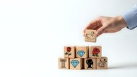Hand placing wooden block with lightbulb icon on stack of cubes featuring various symbols representing creativity, innovation and business concepts.