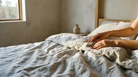 Woman with medium skin tone arranging natural linen bedding on bed in minimalist bedroom with concrete walls. Suitable for home decor, lifestyle, and interior design content.