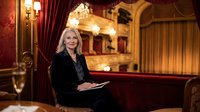 Elegant blonde businesswoman in black suit sitting in luxurious opera house theater box with red velvet curtains and golden interior for corporate events.