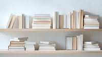 Books arranged on wooden shelves against white wall in minimalist style. Collection of hardcover and paperback volumes in neutral beige and white colors creates calm atmosphere.