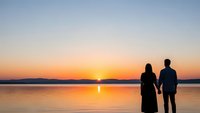 Romantic couple silhouettes holding hands watching golden sunset over calm lake with mountain backdrop creating peaceful evening atmosphere.