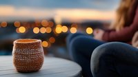 Cozy knitted candle holder with warm glowing light on wooden table against blurred city lights background creates romantic evening atmosphere.