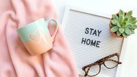 Cozy flat lay with stay home letter board, pastel mug, pink blanket, glasses, and succulent plant on white background.