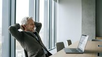 Senior businessman relaxing at modern office desk with laptop, hands behind head in satisfied pose by large windows.