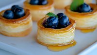 Mini puff pastry tarts filled with honey and topped with fresh blueberries and mint leaves on white plate, sweet dessert close up view.