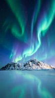 Spectacular aurora borealis dancing over snow-capped mountain peak with vibrant green and purple lights reflecting on calm water surface under starry night sky.