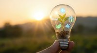 Hand holding light bulb with green plant and renewable energy icons at sunset representing sustainable innovation and eco-friendly technology solutions.