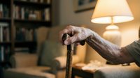 Elderly hands gripping wooden walking cane in warm home interior with lamp and bookshelf background for healthcare and aging concepts.