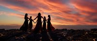 Group of women holding hands in circle silhouetted against dramatic orange sunset sky, spiritual gathering ritual ceremony on rocky landscape.