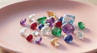 Collection of colorful precious gemstones including diamonds, emeralds, rubies, sapphires and amethysts displayed on pink ceramic plate for jewelry making.