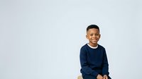 Young boy, approximately 6-8 years old, African descent, wearing dark navy long-sleeve shirt, smiling at camera against white background. Suitable for family, education, and children's lifestyle content.