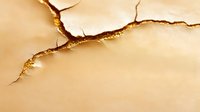 Dramatic crack with golden edges on beige textured surface creates abstract pattern. Natural light emphasizes depth and shadows in minimalist composition.
