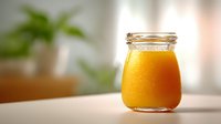 Fresh orange juice in glass jar with condensation droplets on bright table, blurred green plant background for healthy breakfast concept.