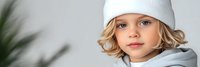 Young blonde Caucasian girl wearing white knit beanie hat and gray hoodie looking at camera with serious expression in modern indoor setting.