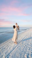 Young Caucasian mother in white dress lifting happy baby on white sand beach at pastel sunset, joyful family moment by the sea.