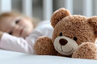 Soft brown teddy bear in foreground with sleeping child blurred in background on white bedding, creating peaceful naptime atmosphere. Childhood comfort concept.