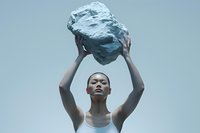 Asian woman in white tank top holding large stone above head against light blue background, expressing strength and determination in minimalist setting.