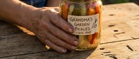 Elderly hands holding homemade pickled vegetables jar labeled Grandmas Garden Pickles on rustic wooden table for traditional cooking and preservation.