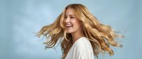 Young blonde Caucasian woman with flowing wavy hair laughing joyfully against soft blue background. Natural beauty and happiness concept for lifestyle content.