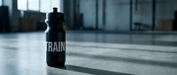 Black sports water bottle with "TRAIN" text on concrete gym floor with natural light from windows, suitable for fitness, wellness, and athletic lifestyle marketing.